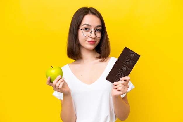 Young Ukrainian woman isolated on yellow background taking a chocolate tablet in one hand and an apple in the other