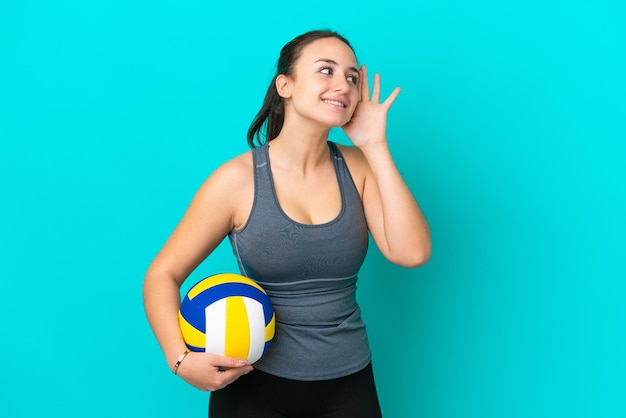 Young Ukrainian woman playing volleyball isolated on blue background listening to something by putting hand on the ear