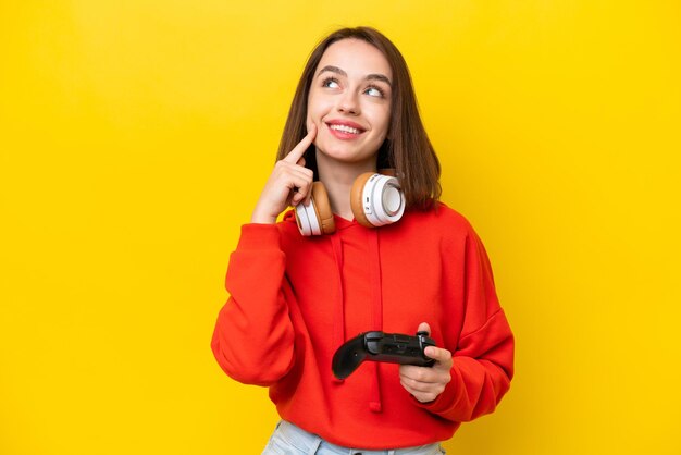 Young ukrainian woman playing with a video game controller isolated on yellow background thinking an idea while looking up