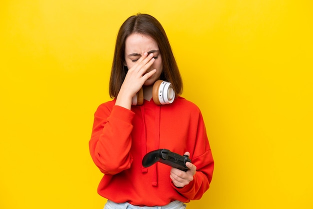 Young Ukrainian woman playing with a video game controller isolated on yellow background with tired and sick expression