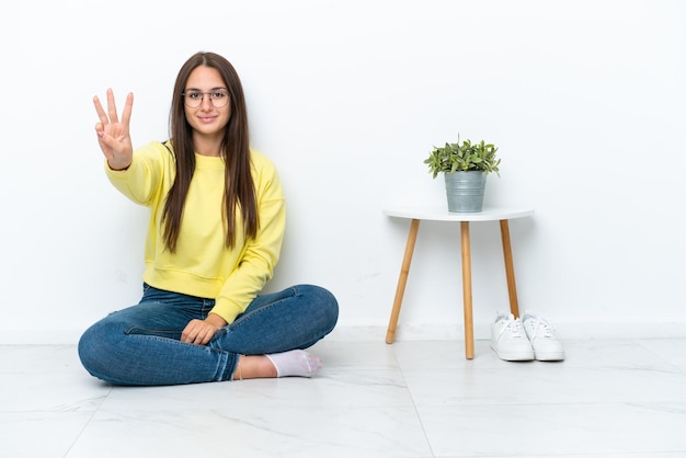 Giovane donna ucraina seduta sul pavimento della sua casa isolata sul muro bianco felice e contando tre con le dita Foto Premium