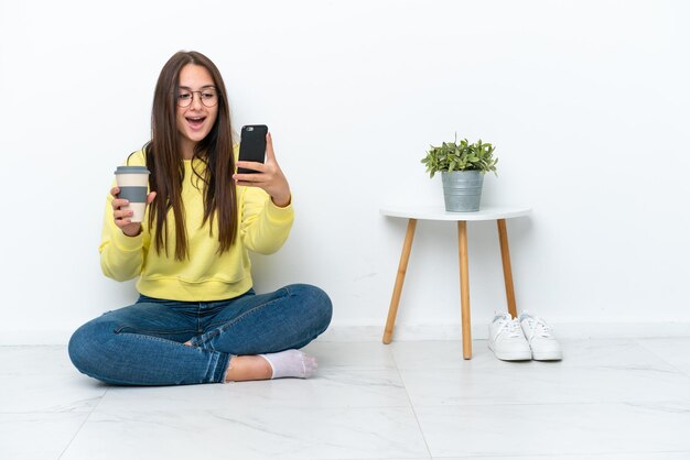 Young Ukrainian woman sitting on the floor of her house isolated on white wall holding coffee to take away and a mobile