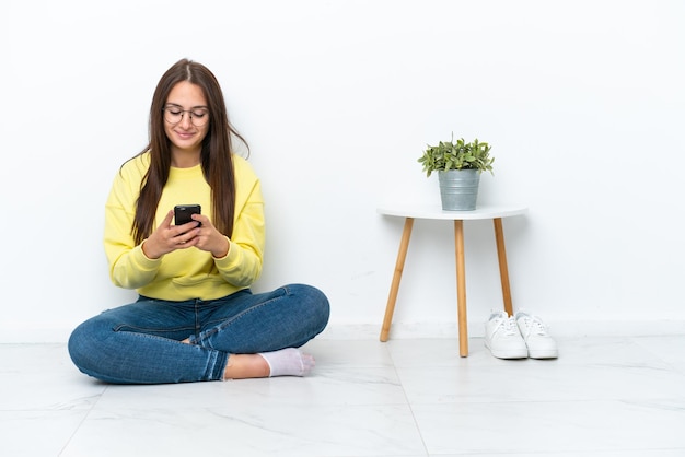 Young Ukrainian woman sitting on the floor of her house isolated on white wall sending a message with the mobile