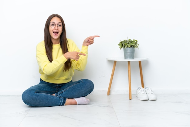 Young Ukrainian woman sitting on the floor of her house isolated on white wall surprised and pointing side