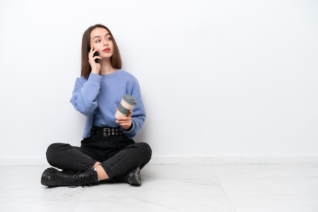 Young Ukrainian woman sitting on the floor isolated on white background holding coffee to take away and a mobile