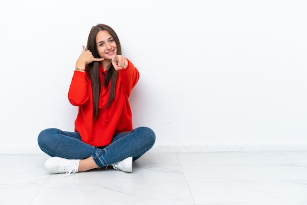 Young Ukrainian woman sitting on the floor isolated on white background making phone gesture and pointing front