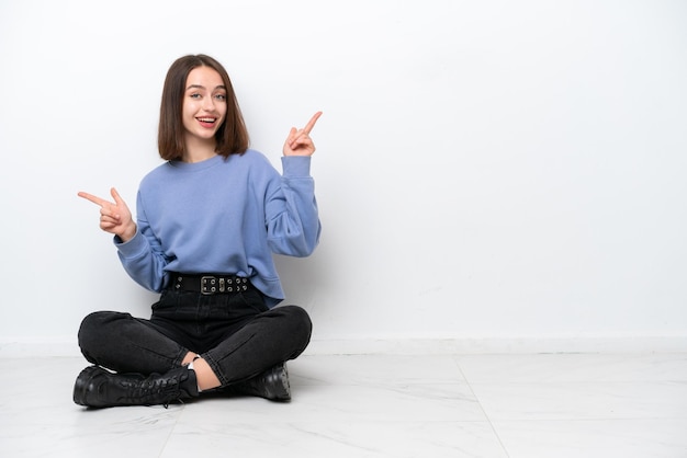 Young Ukrainian woman sitting on the floor isolated on white background pointing finger to the laterals and happy