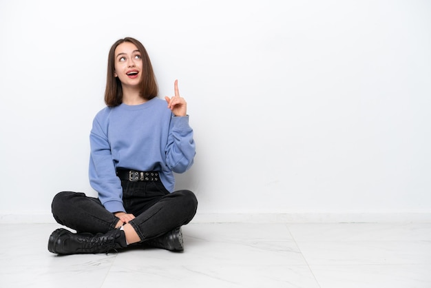 Young Ukrainian woman sitting on the floor isolated on white background pointing up and surprised