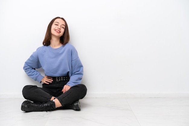 Young Ukrainian woman sitting on the floor isolated on white background posing with arms at hip and smiling