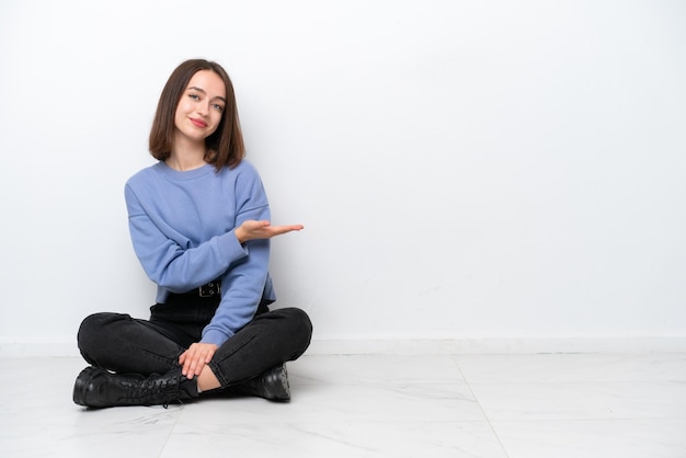 Young Ukrainian woman sitting on the floor isolated on white background presenting an idea while looking smiling towards