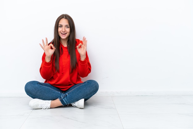 Young Ukrainian woman sitting on the floor isolated on white background showing an ok sign with fingers