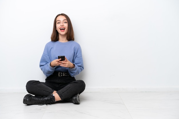 Young Ukrainian woman sitting on the floor isolated on white background surprised and sending a message