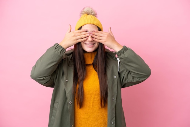 Young Ukrainian woman wearing winter jacket isolated on pink background covering eyes by hands