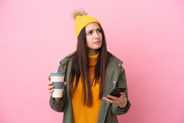 持ち帰り用のコーヒーと何かを考えながら携帯電話を保持しているピンクの背景に分離された冬のジャケットを着ている若いウクライナの女性