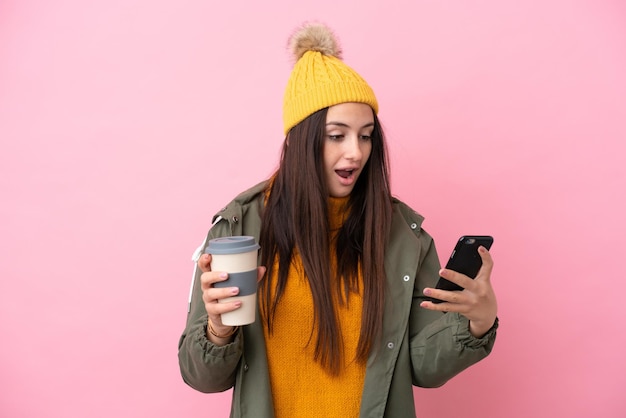 Young ukrainian woman wearing winter jacket isolated on pink
background holding coffee to take away and a mobile