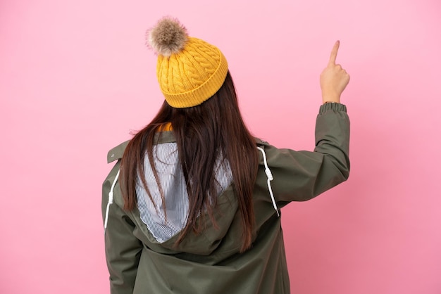 Young Ukrainian woman wearing winter jacket isolated on pink background pointing back with the index finger