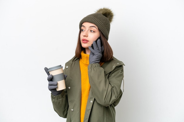 Молодая украинка в зимней куртке на белом фоне держит кофе на вынос и мобильный телефон