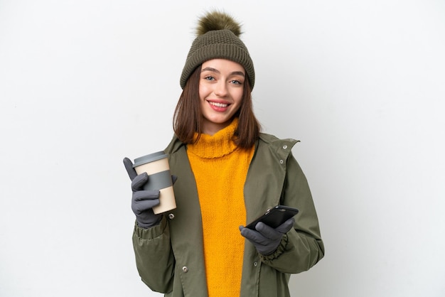 Молодая украинка в зимней куртке на белом фоне держит кофе на вынос и мобильный телефон