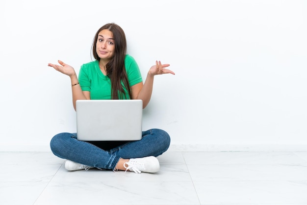 Young Ukrainian woman with a laptop sitting on floor having doubts while raising hands