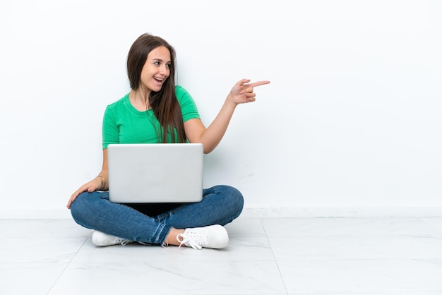 Young Ukrainian woman with a laptop sitting on floor pointing finger to the side and presenting a product