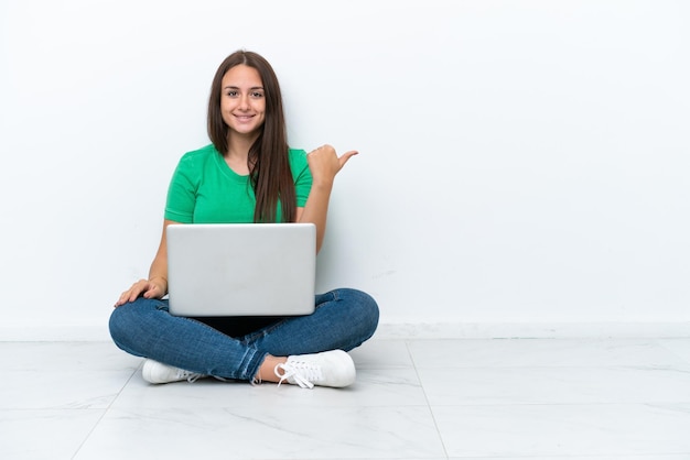 Young Ukrainian woman with a laptop sitting on floor pointing to the side to present a product