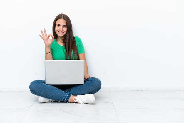 Young Ukrainian woman with a laptop sitting on floor showing ok sign with fingers