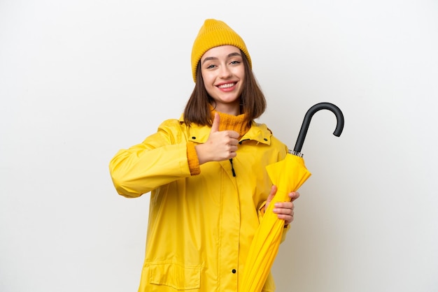 防雨コートと親指のジェスチャーを与える白い背景で隔離の傘を持つ若いウクライナ人女性