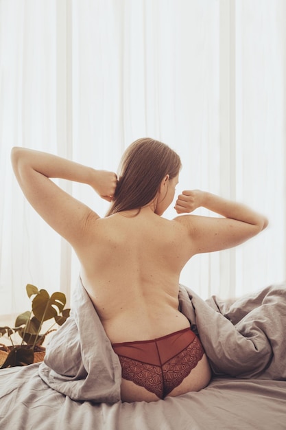Молодая раздетая женщина сидит на кровати с серым постельным бельем