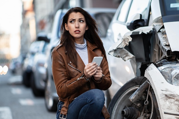 Giovane donna sfortunata che utilizza smartphone e messaggi di testo dopo un incidente d'auto in città