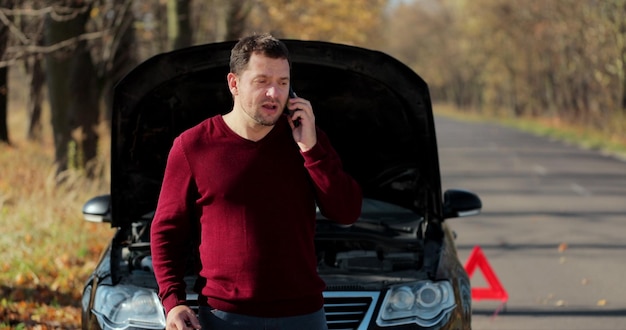 Young unhappy man using smartphone calling car assistance services standing by breakdown automobile on the roadside. Helpless man need help and assistance.