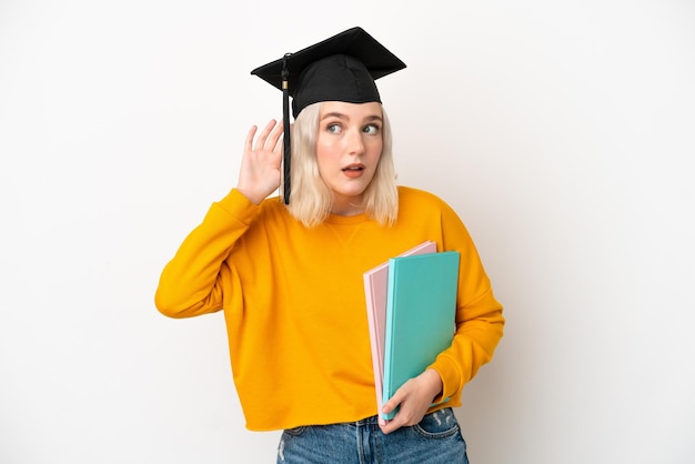 Young university caucasian woman graduate isolated on white background listening to something by putting hand on the ear