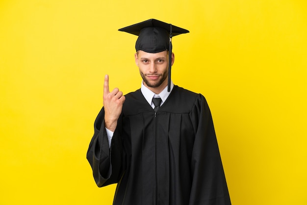 人差し指で指している黄色の背景に分離された若い大学卒業生の白人男性素晴らしいアイデア