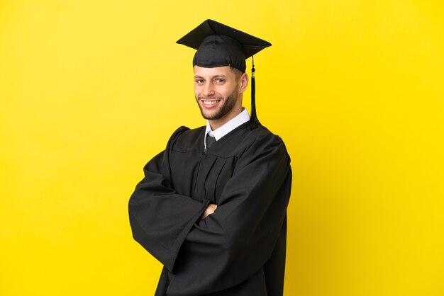 腕を組んで、楽しみにして黄色の背景に分離された若い大学卒業生の白人男性