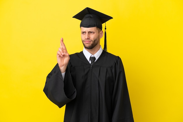 指が交差し、最高を願って黄色の背景に分離された若い大学卒業生の白人男性