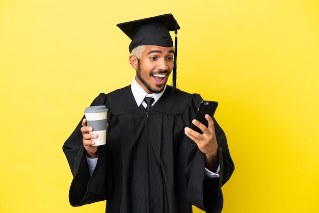 Young university graduate Colombian man isolated on yellow background holding coffee to take away and a mobile