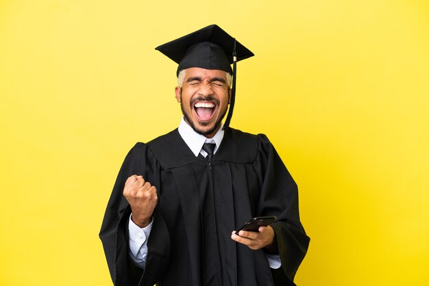 Young university graduate Colombian man isolated on yellow background with phone in victory position