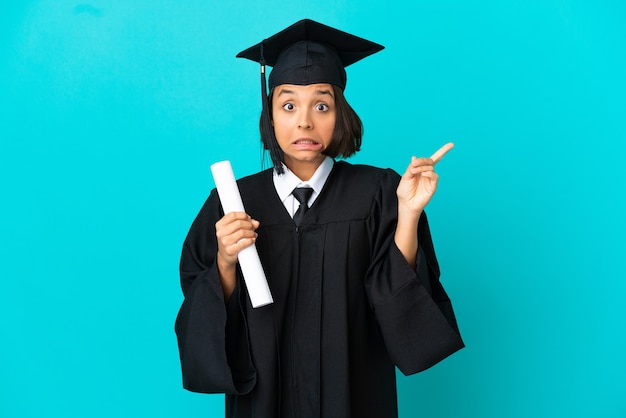 Young university graduate girl over isolated blue background frightened and pointing to the side