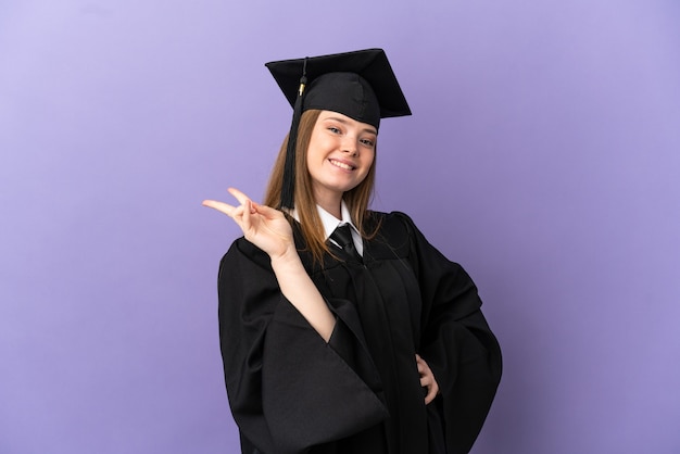 Young university graduate over isolated purple background smiling and showing victory sign