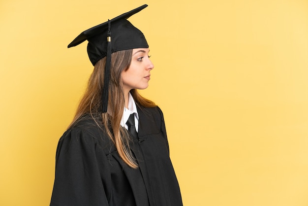 Young university graduate isolated on yellow background looking to the side