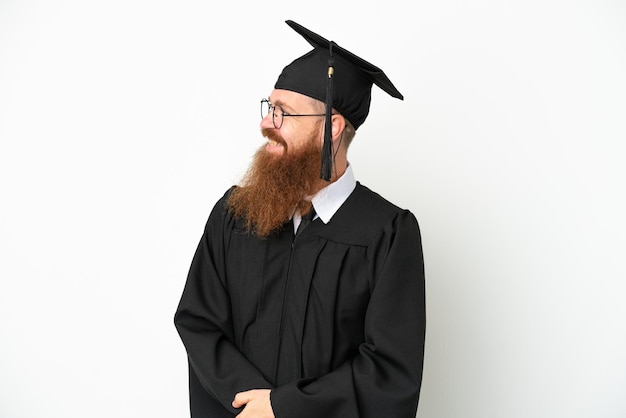 横を見て笑っている白い背景に分離された若い大学卒業生の赤みを帯びた男