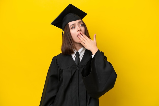 あくびをし、手で大きく開いた口を覆う黄色の背景に分離された若い大学卒業生ウクライナの女性
