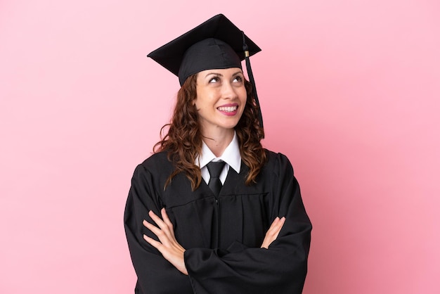 Young university graduate woman isolated on pink background\
looking up while smiling