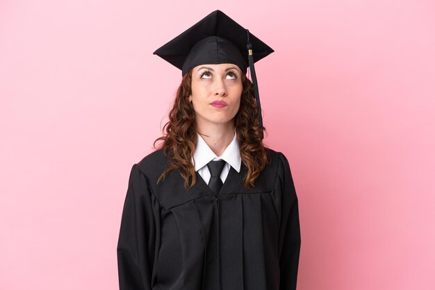 Young university graduate woman isolated on pink background and\
looking up