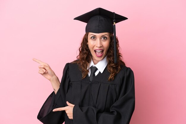 Young university graduate woman isolated on pink background surprised and pointing side