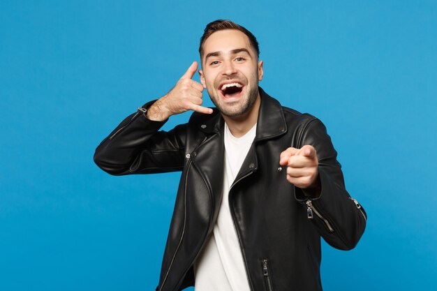 Young unshaven man in black jacket white t-shirt looking camera doing phone gesture like says call me back isolated on blue wall background studio portrait. People lifestyle concept Mock up copy space