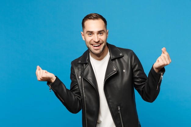 Young unshaven man in black jacket white t-shirt rubbing fingers, showing cash gesture, asking for money isolated on blue wall background studio portrait. People lifestyle concept. Mock up copy space.