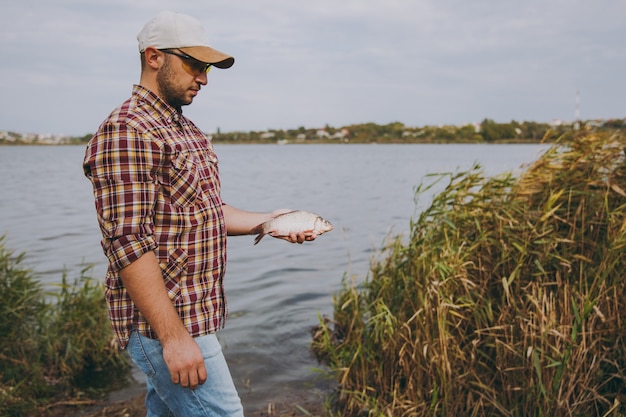 Young unshaven man in checkered shirt, cap and sunglasses caught a fish and holds it in arms on shore of lake on background of water, shrubs and reeds. Lifestyle, fisherman recreation, leisure concept