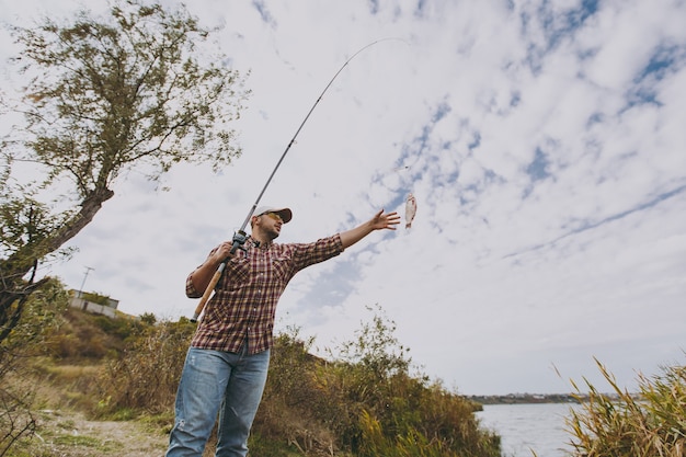 Young unshaven man in checkered shirt, cap, sunglasses holds fishing pole and extends his hand to caught fish on shore of lake near shrubs and reeds. lifestyle, recreation, fisherman leisure concept