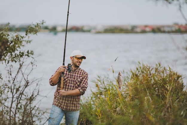 Young unshaven man in checkered shirt, cap and sunglasses pulled out fishing pole with caught fish and rejoices on shore of lake near shrubs and reeds. Lifestyle, recreation, fisherman leisure concept