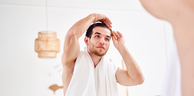 Young unshaven man combing hair standing near mirror in bathroom